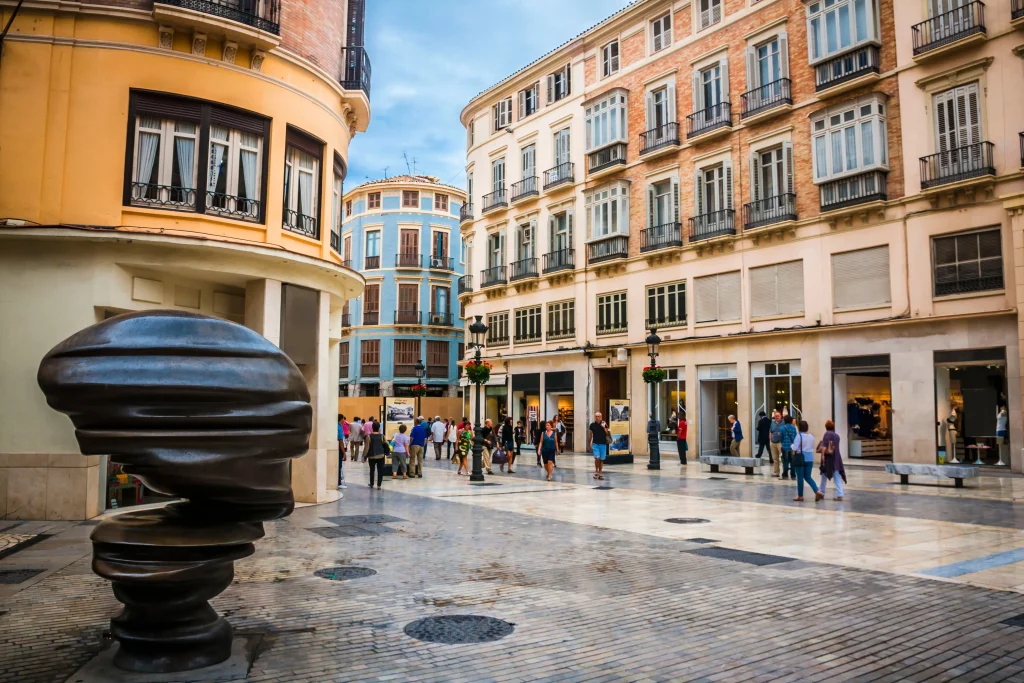 Plaza con edificios hist&oacute;ricos y escultura moderna, gente paseando.