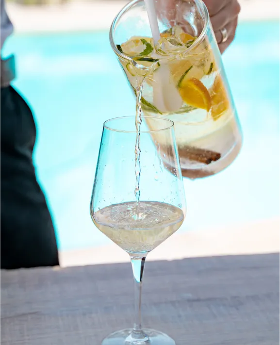 Pessoa servindo bebida refrescante com frutas em ta&ccedil;a com piscina ao fundo.