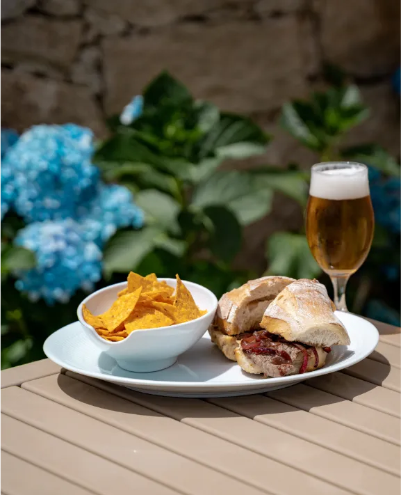 Sandu&iacute;che e nachos com cerveja ao ar livre, flores azuis ao fundo.