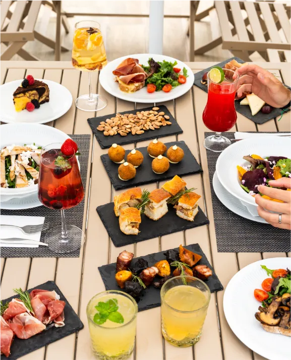 Mesa com pratos diversos, saladas e bebidas coloridas ao ar livre.