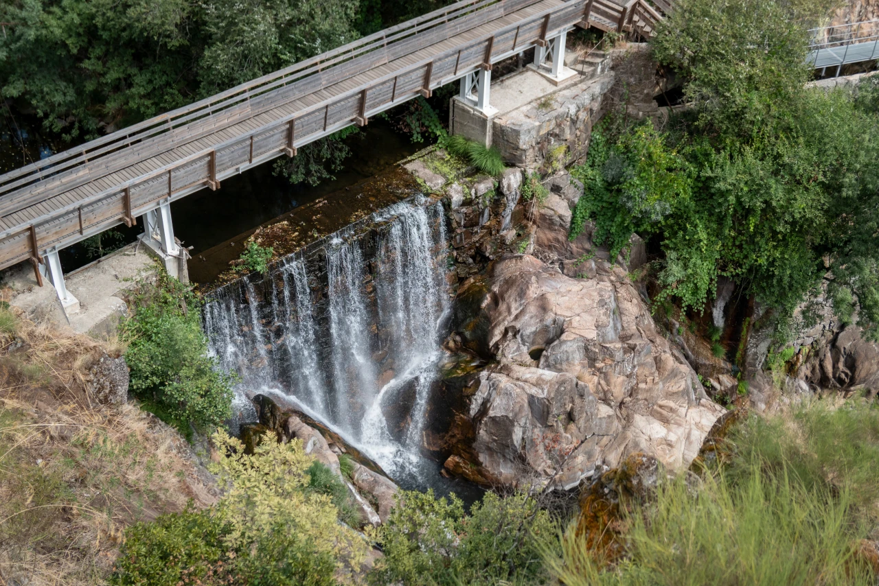 Ponte de madeira sobre cascata em meio a rochas e vegeta&ccedil;&atilde;o densa.