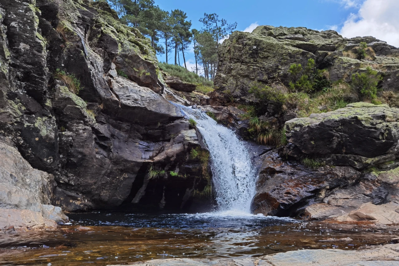 Cachoeira entre rochas com &aacute;rvores ao fundo e c&eacute;u azul.