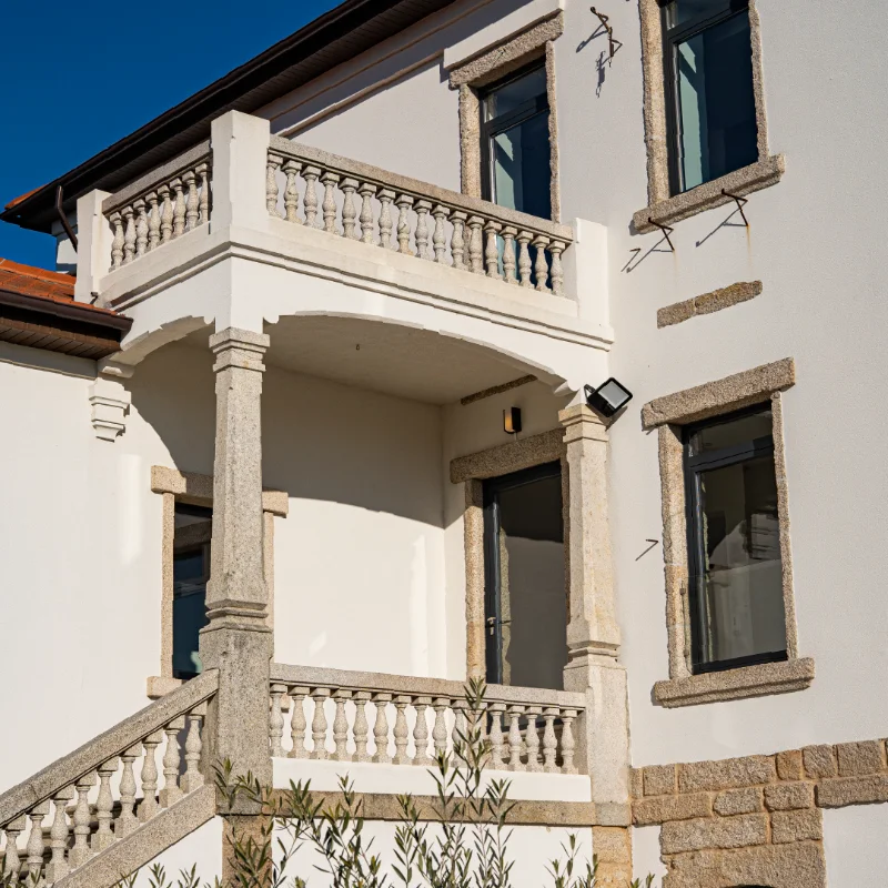 Fachada de casa com varanda e pilastras de pedra sob c&eacute;u azul.