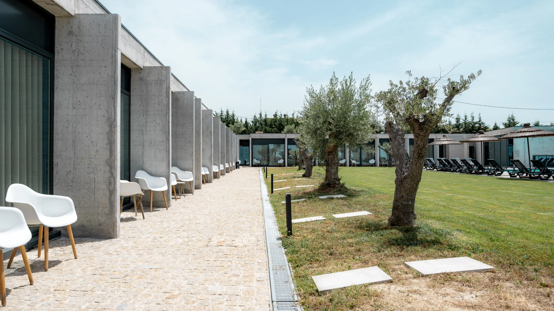 Corredor com cadeiras brancas ao lado de gramado e &aacute;rvores; c&eacute;u claro.
