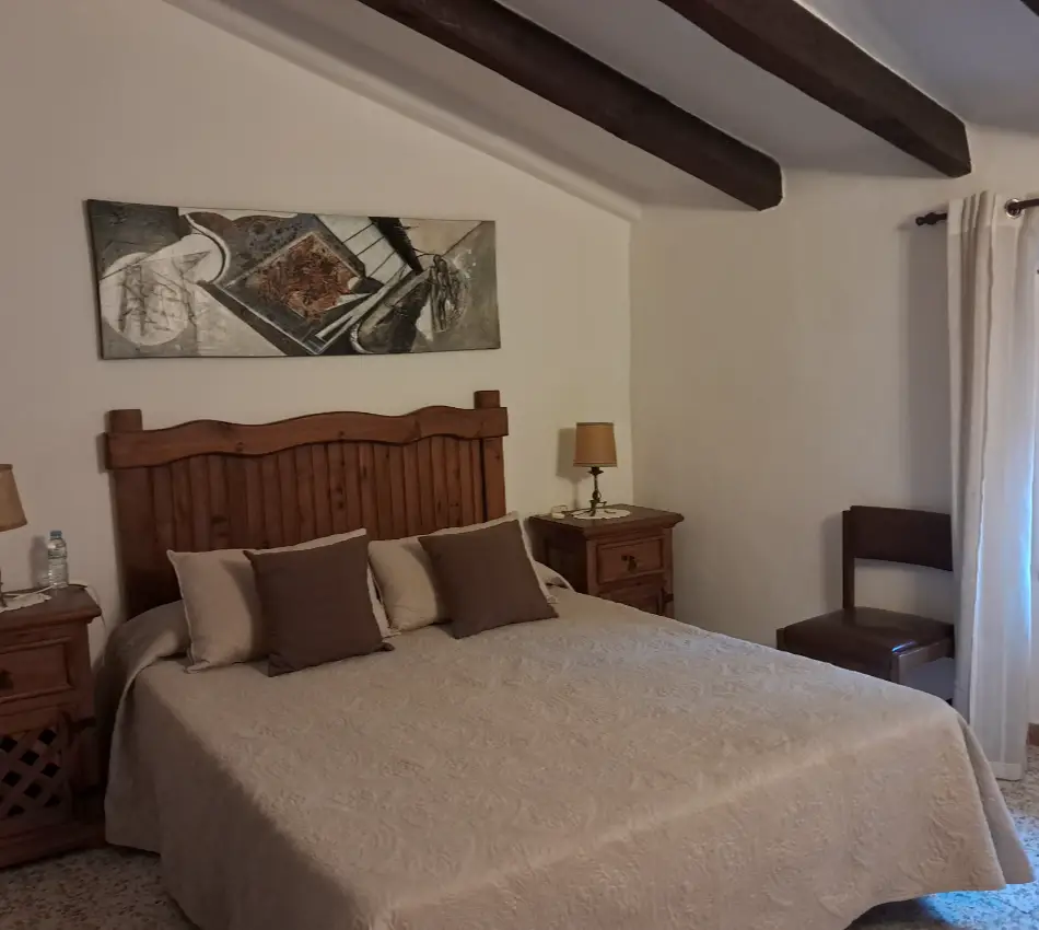 Dormitori amb sostre de bigues, llit doble i tauletes de nit de fusta.