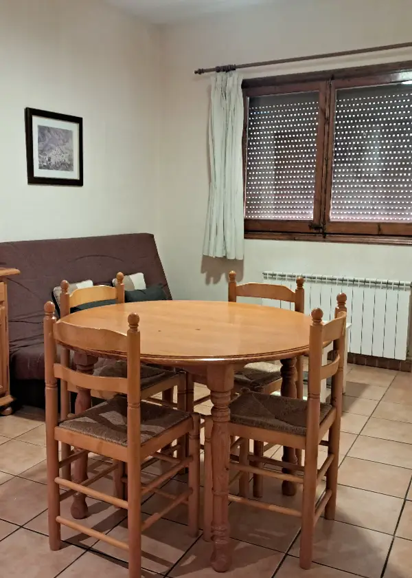 Sala d'estar amb taula rodona i cadires de fusta, sof&agrave; i finestra amb cortina.