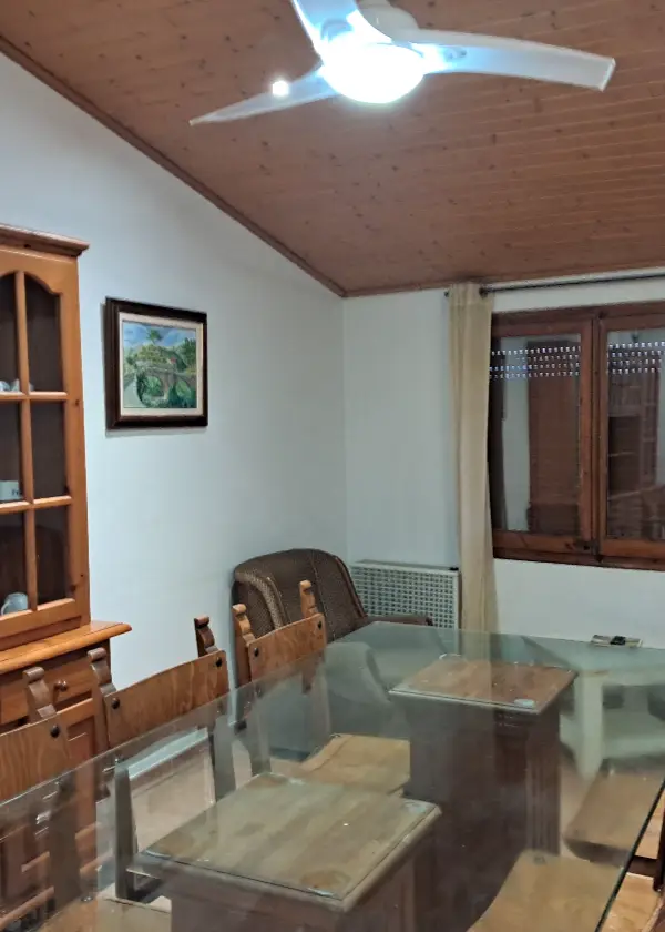 Sala d'estar amb taula de vidre, cadires de fusta, i ventilador de sostre.