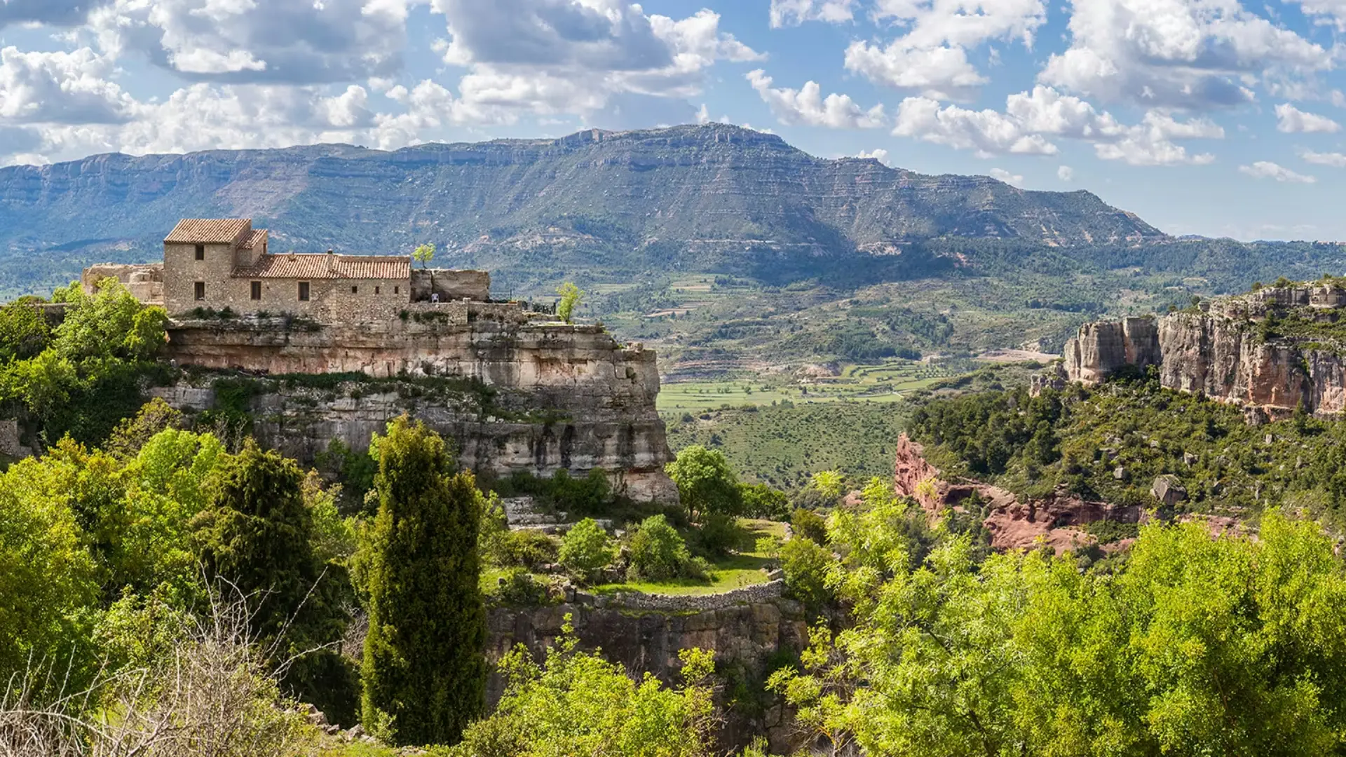 Paisatge amb muntanyes, una casa de pedra al penya-segat i arbres verds.