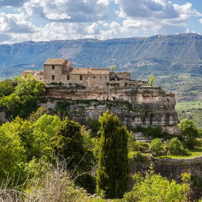 Casa de pedra al cim d'un penya-segat amb muntanyes al fons.