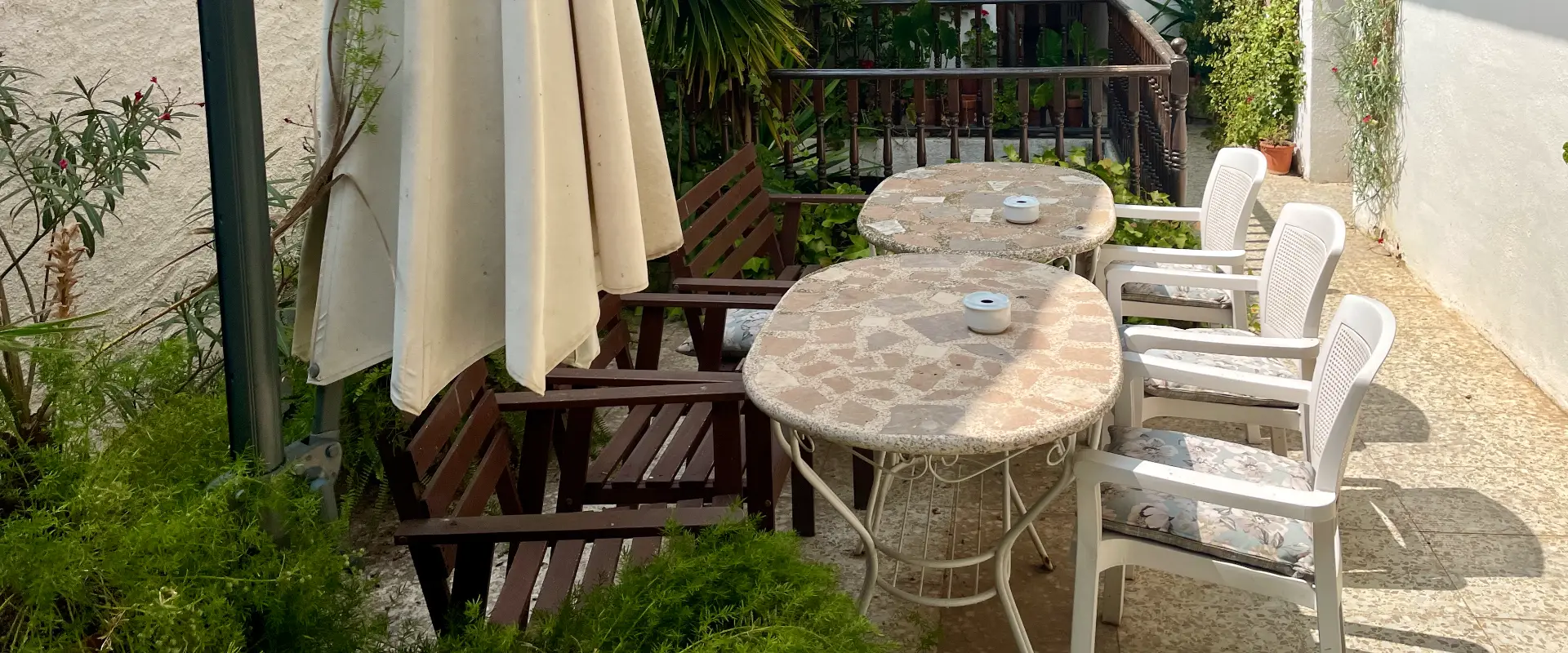 Taules i cadires exteriors amb para-sol en terrassa envoltada de plantes.