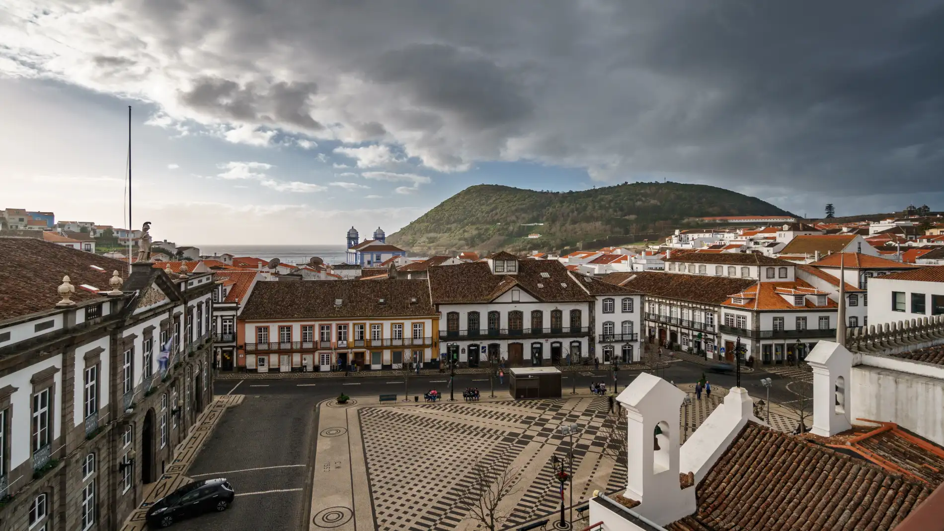 Pra&ccedil;a com edif&iacute;cios antigos, fundo montanhoso e c&eacute;u nublado.