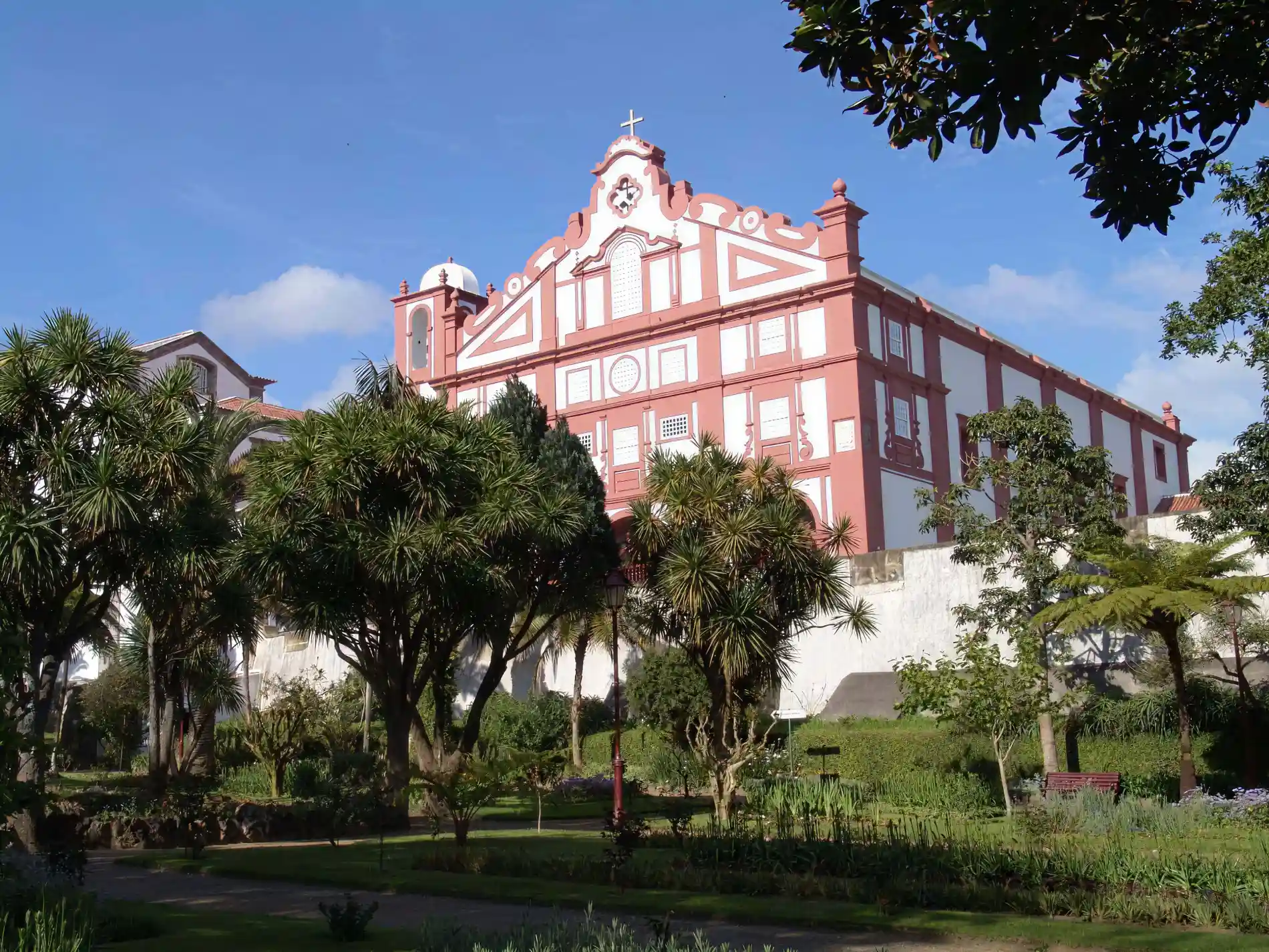 Edif&iacute;cio hist&oacute;rico rosa e branco rodeado por &aacute;rvores e jardim ao ar livre.