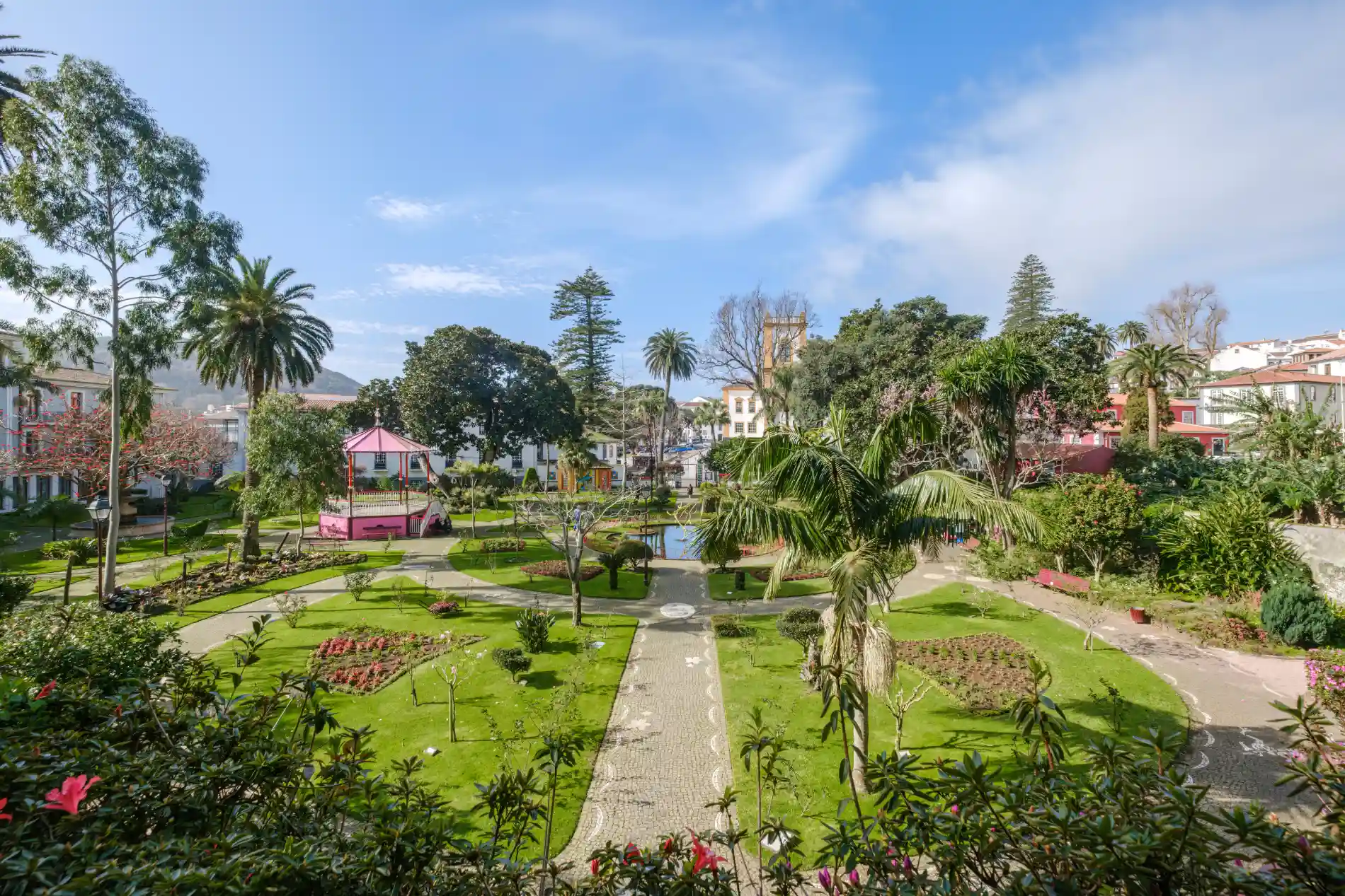 Parque com jardim verde, caminhos e coreto sob c&eacute;u azul.