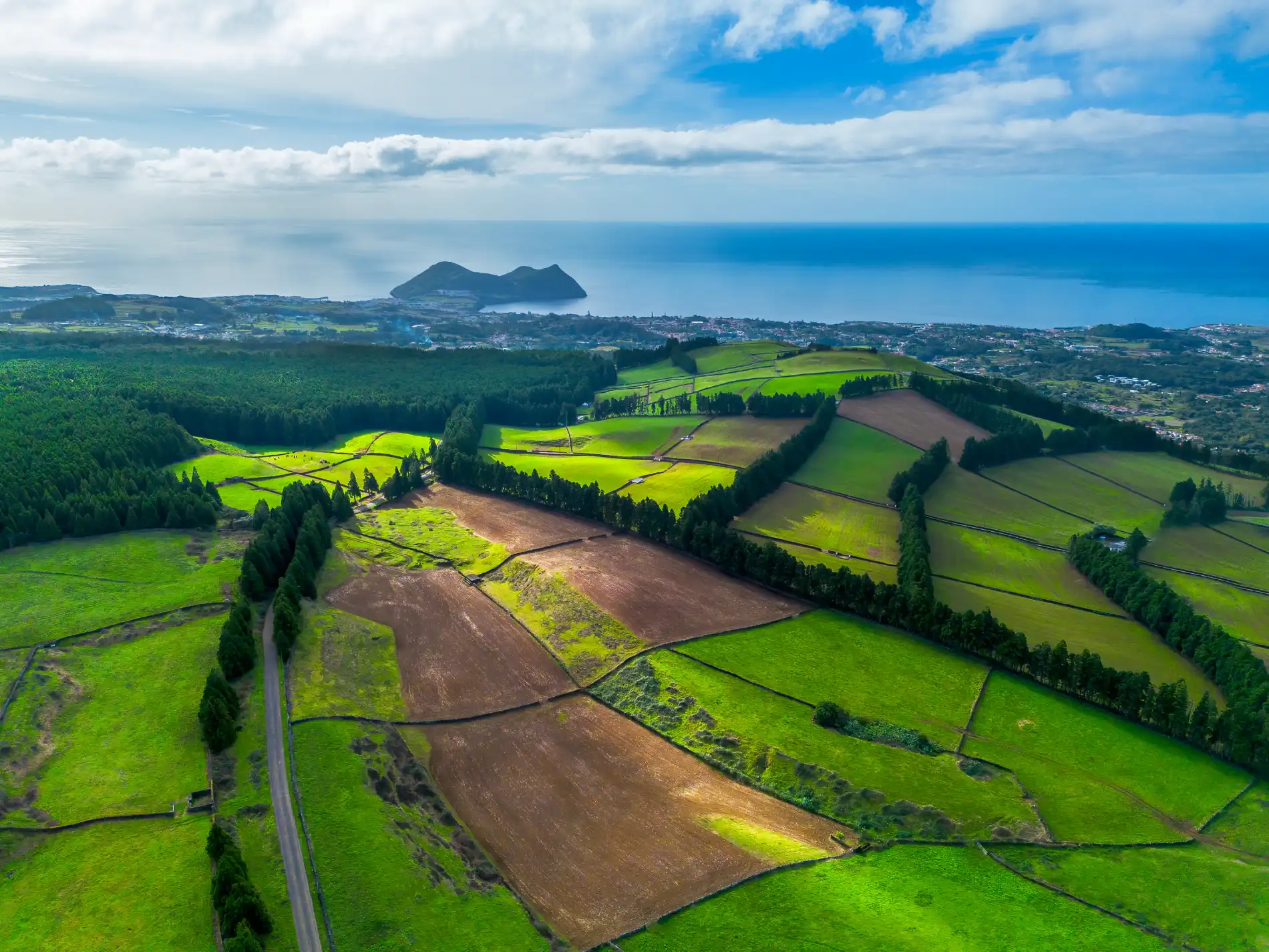 Paisagem a&eacute;rea com campos verdes e mar ao fundo sob c&eacute;u azul.