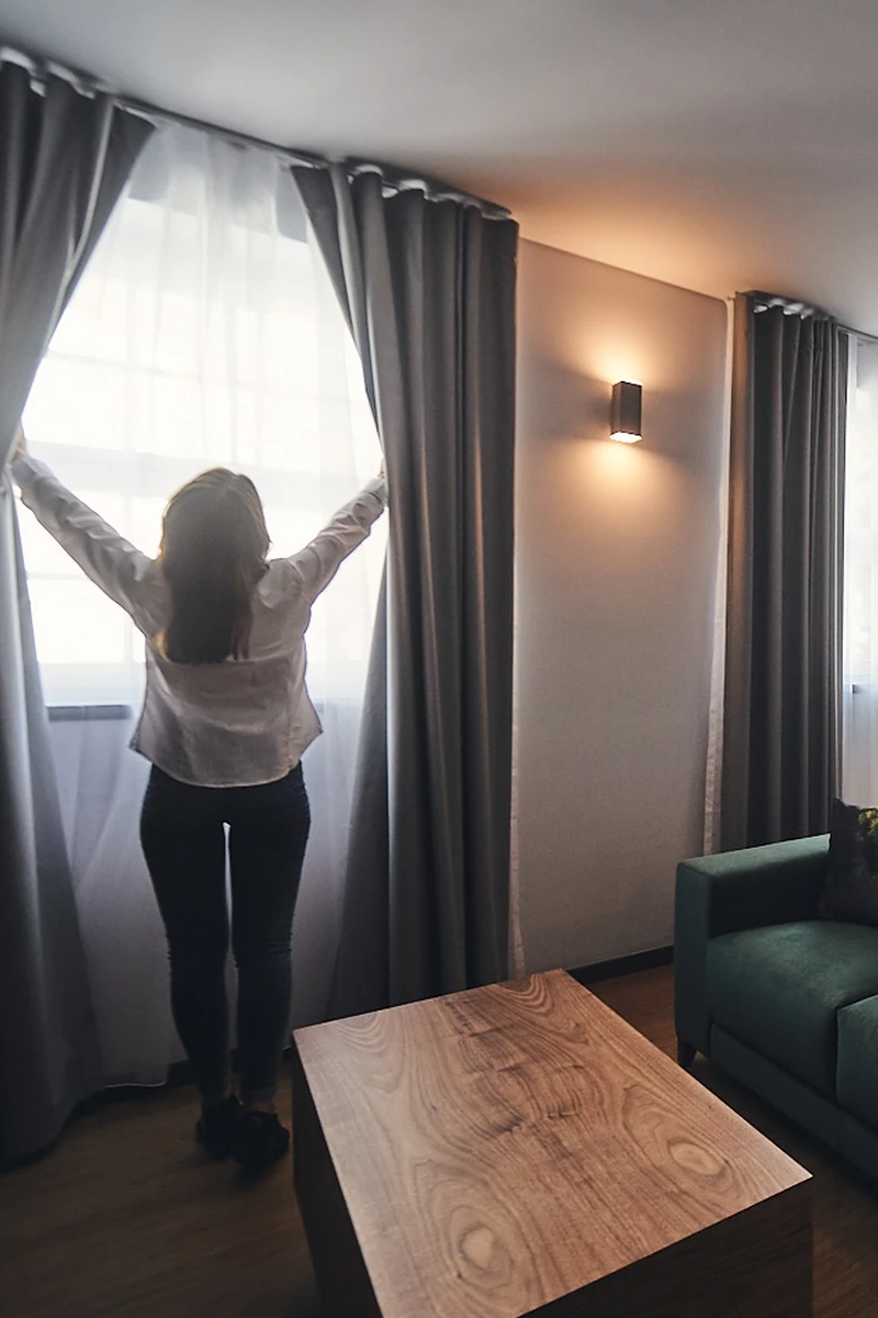 Pessoa abrindo cortina em sala iluminada; sof&aacute; e mesa ao lado.