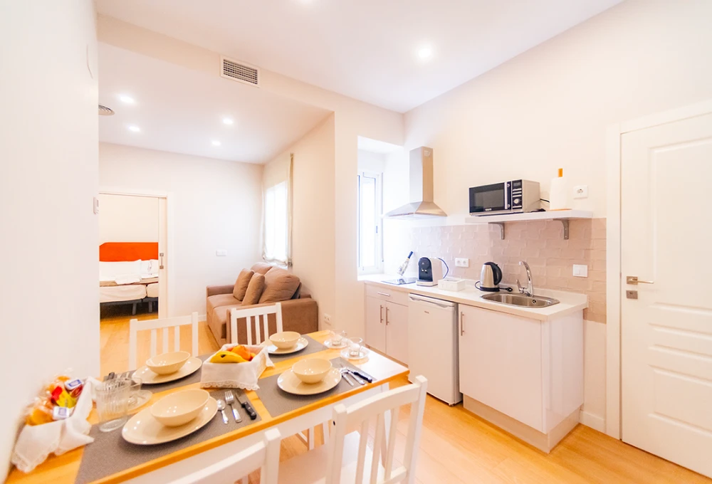 Cocina y comedor modernos con sof&aacute; y dormitorio al fondo, vajilla en la mesa y electrodom&eacute;sticos.