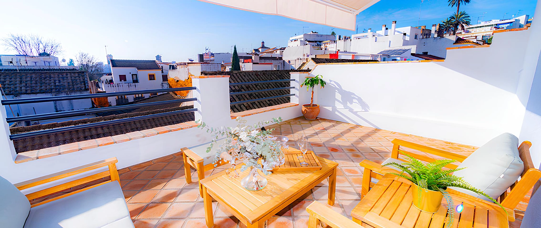 Terraza con muebles de madera y plantas, vista a tejados y cielo despejado.