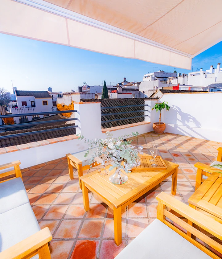 Terraza soleada con muebles de madera y plantas, vista de la ciudad bajo un cielo despejado.