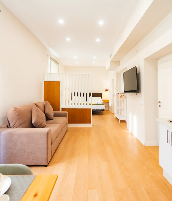 Sala moderna con sof&aacute;, cama al fondo, televisor en la pared y piso de madera clara.