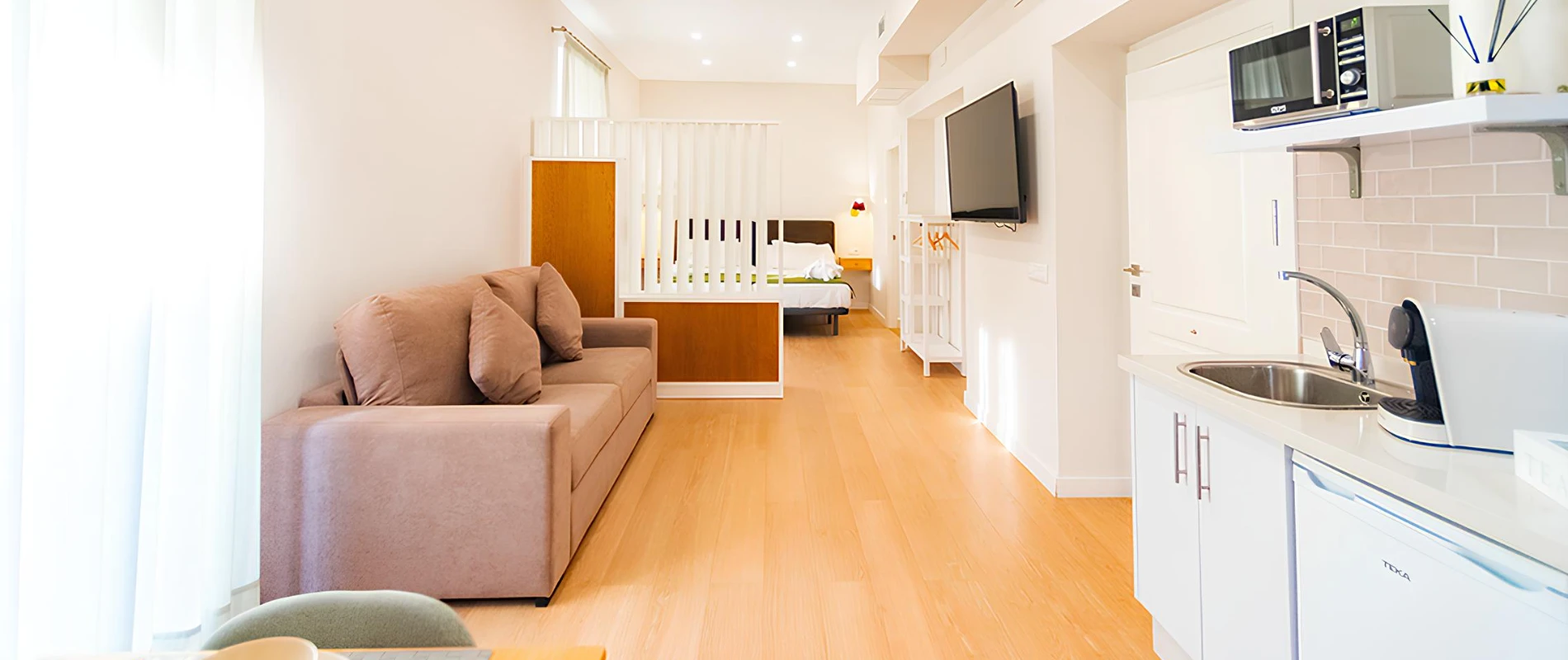 Sala moderna con sof&aacute;, cocina peque&ntilde;a, TV y cama al fondo. Decoraci&oacute;n minimalista y luminosa.