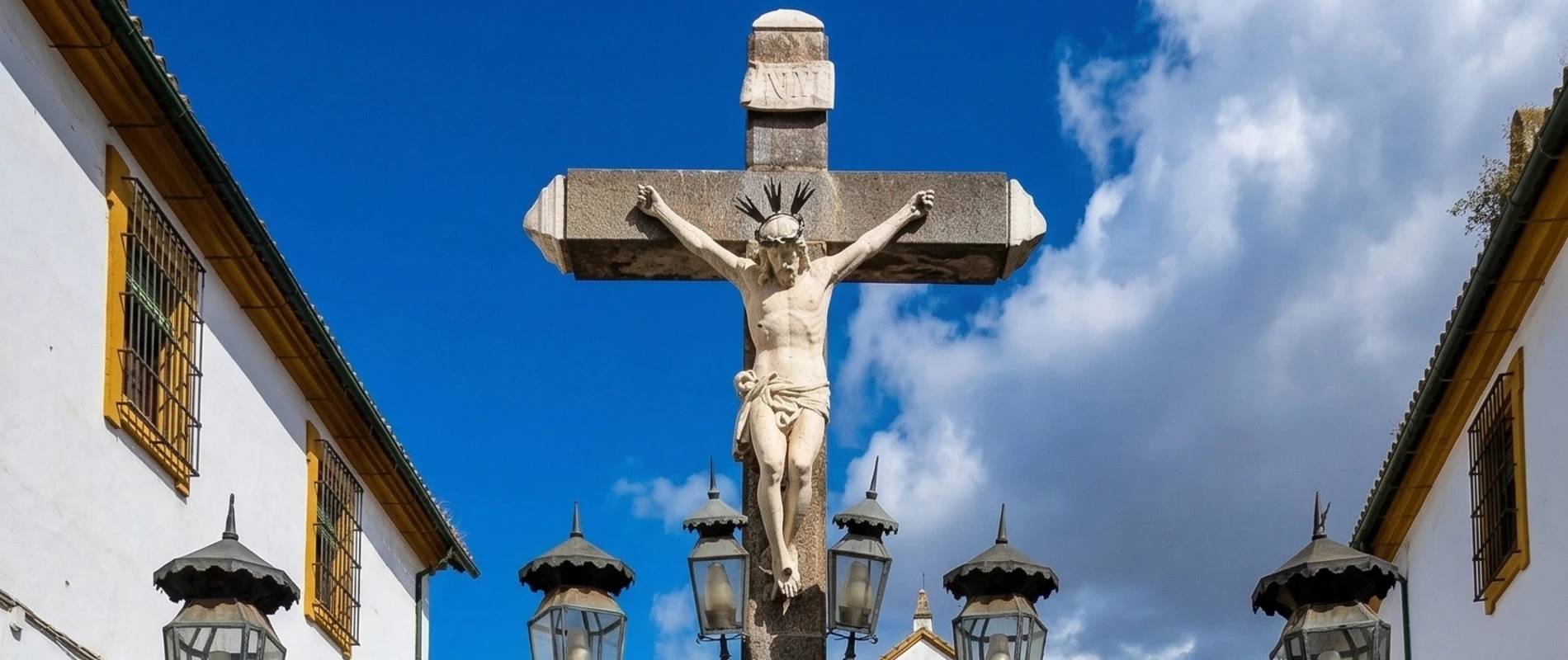 Crucifijo entre dos edificios y farolas, cielo azul con nubes al fondo.