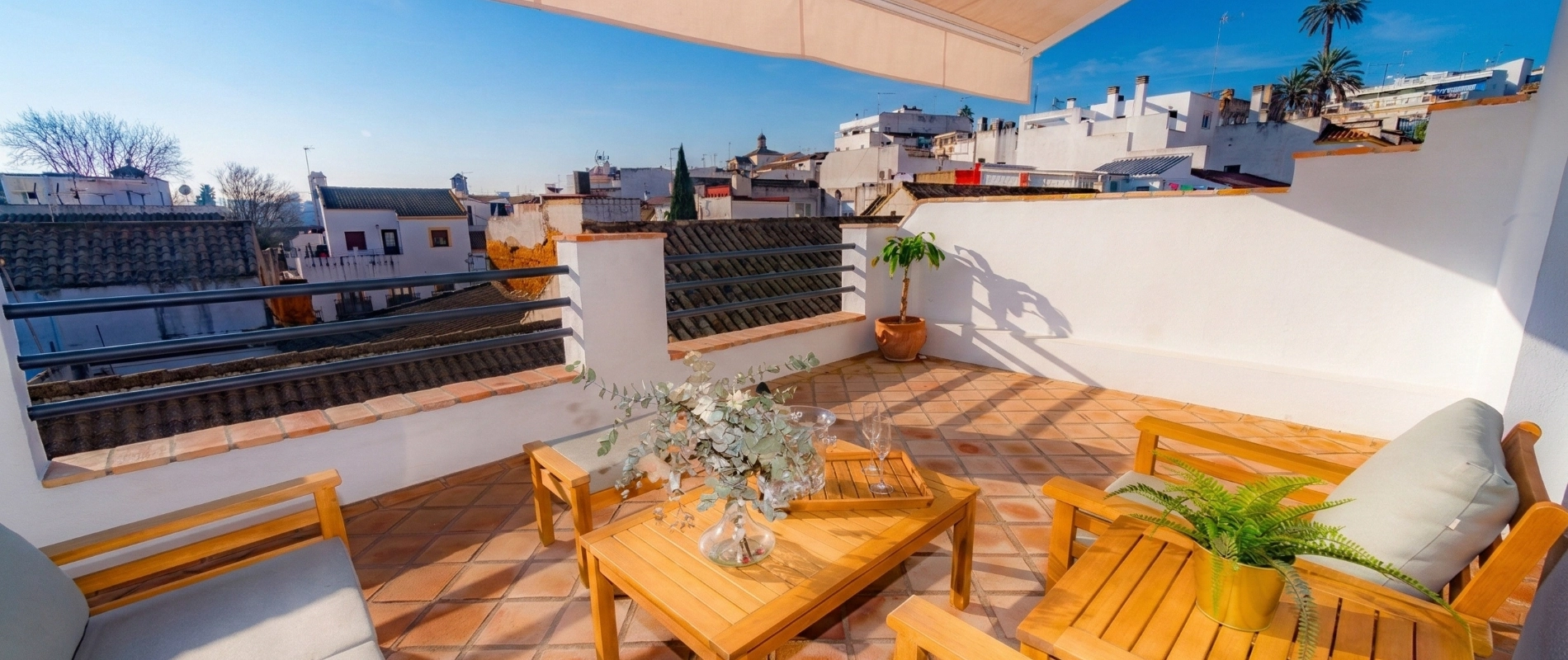 Terraza con muebles de madera, plantas y vista a tejados y cielo despejado.
