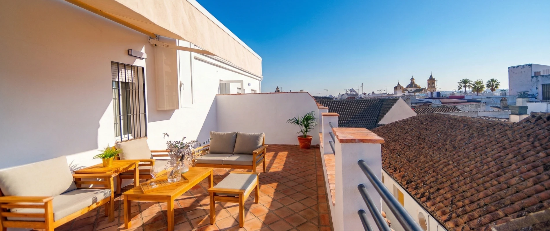 Terraza soleada con muebles de madera, plantas y vistas a tejados y torres bajo un cielo despejado.