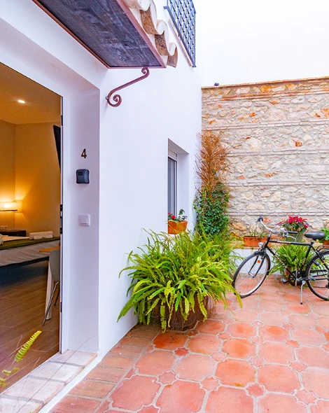 Patio con bicicleta, plantas y puerta abierta; suelo de baldosas y pared de piedra.