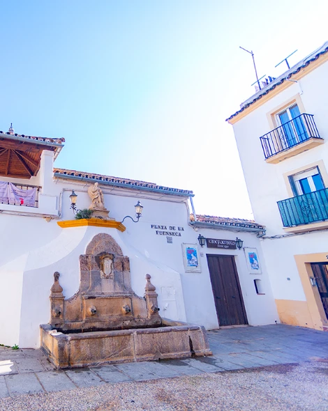 Plaza con fuente hist&oacute;rica y edificios blancos con balcones en un d&iacute;a soleado.