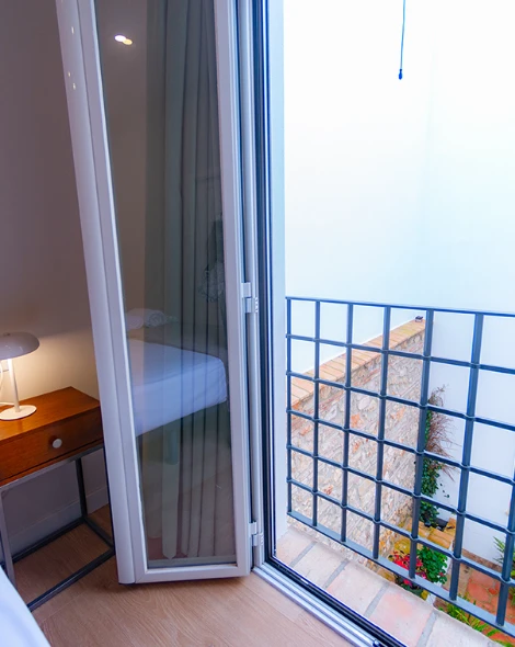 Puerta de cristal abierta, mesa con l&aacute;mpara a la izquierda, barandilla met&aacute;lica afuera.