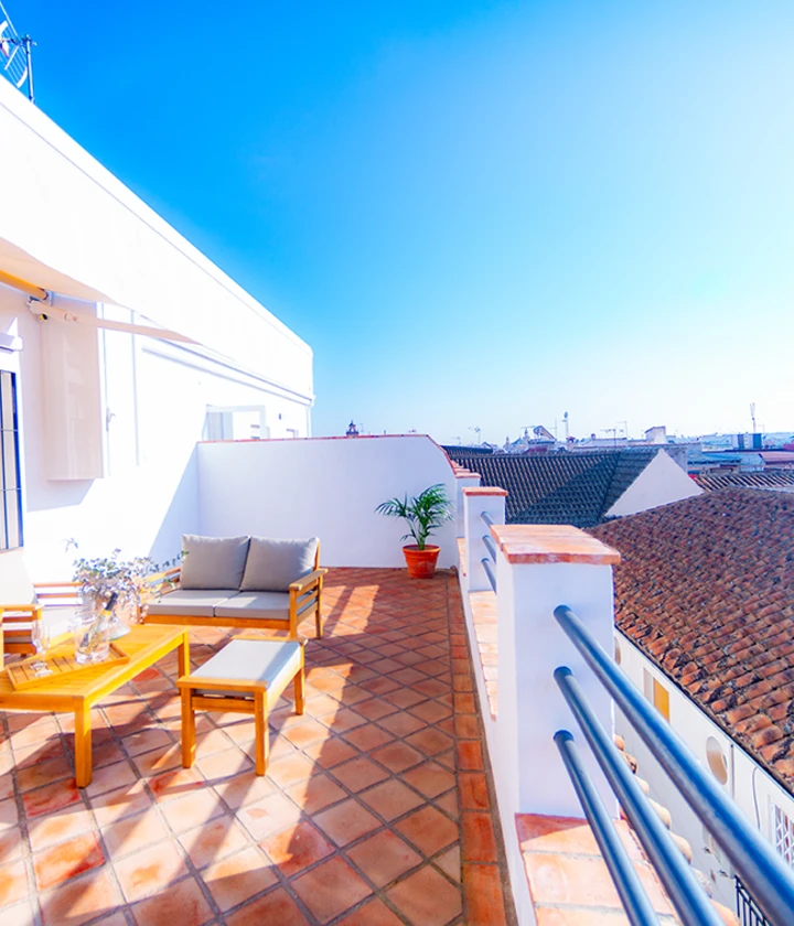 Terraza soleada con muebles y plantas, vista a tejados y cielo despejado.