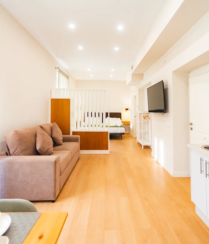 Sala moderna con sof&aacute; marr&oacute;n, cama al fondo y televisor montado en la pared.