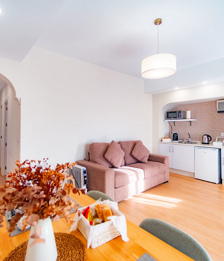 Sala moderna con cocina integrada, sof&aacute; marr&oacute;n y mesa con flores secas sobre mantel trenzado.