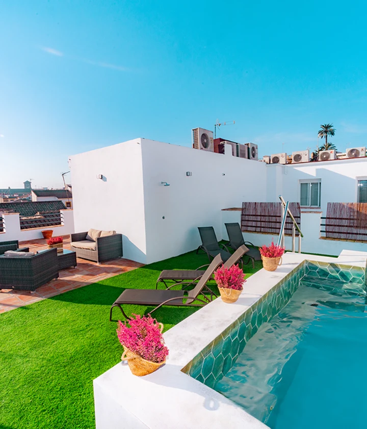 Terraza con piscina, tumbonas, sillones y flores bajo un cielo azul claro.