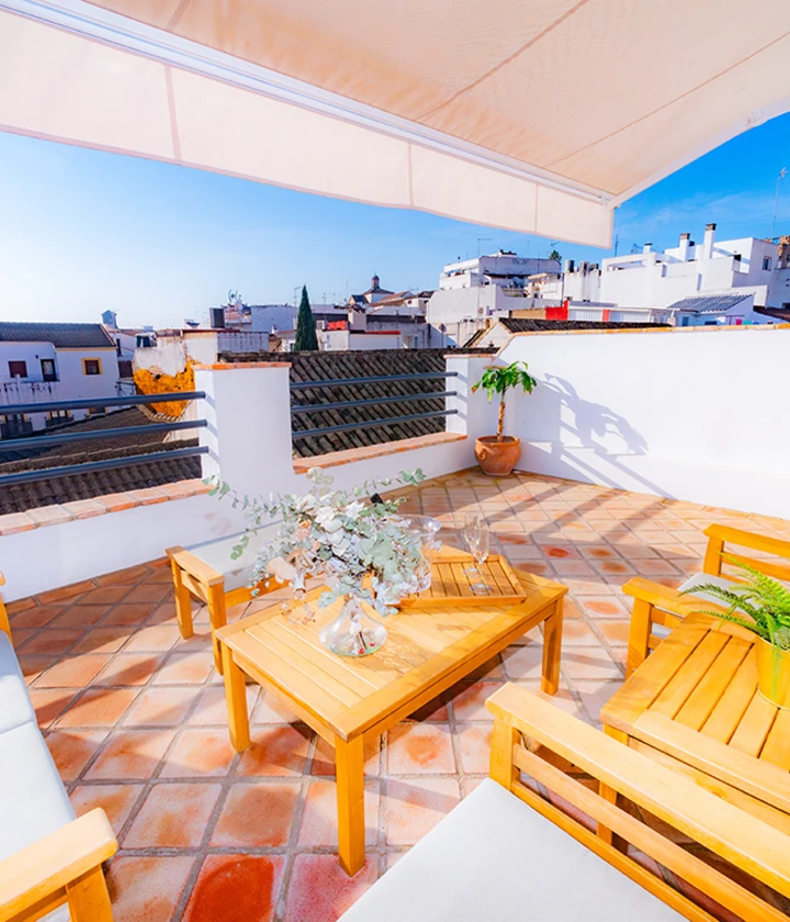 Terraza con muebles de madera y planta en maceta, vista a casas blancas y cielo despejado.