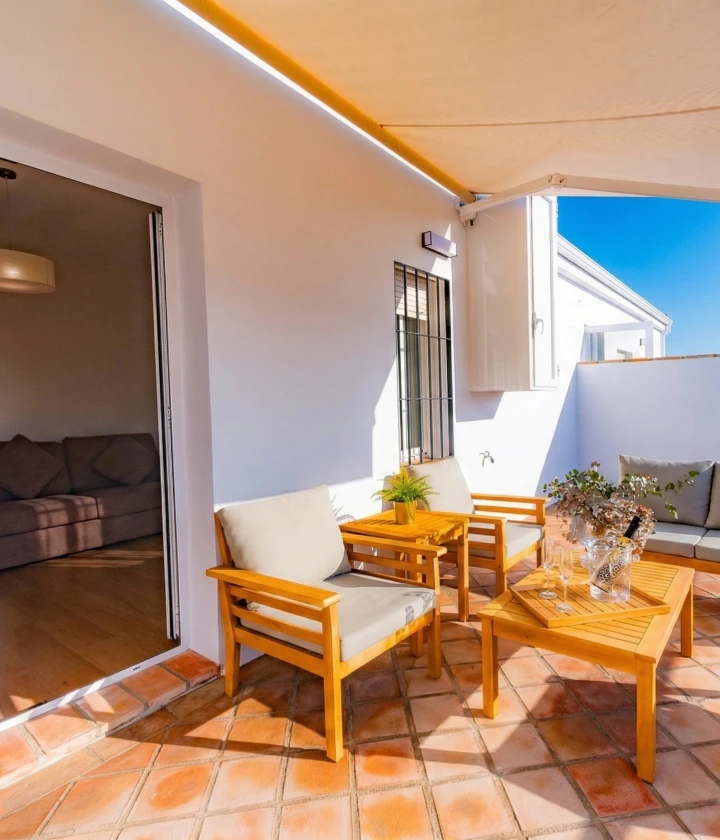 Terraza soleada con sillas y mesa de madera, plantas y sof&aacute; beige, bajo un toldo amarillo.