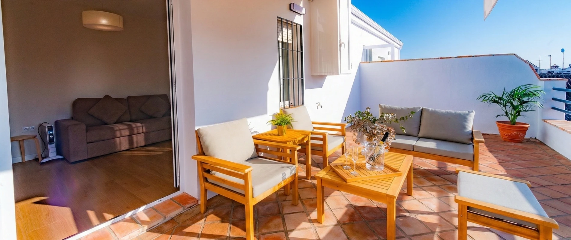 Terraza soleada con muebles de madera, plantas en macetas y puerta corredera a la sala de estar.
