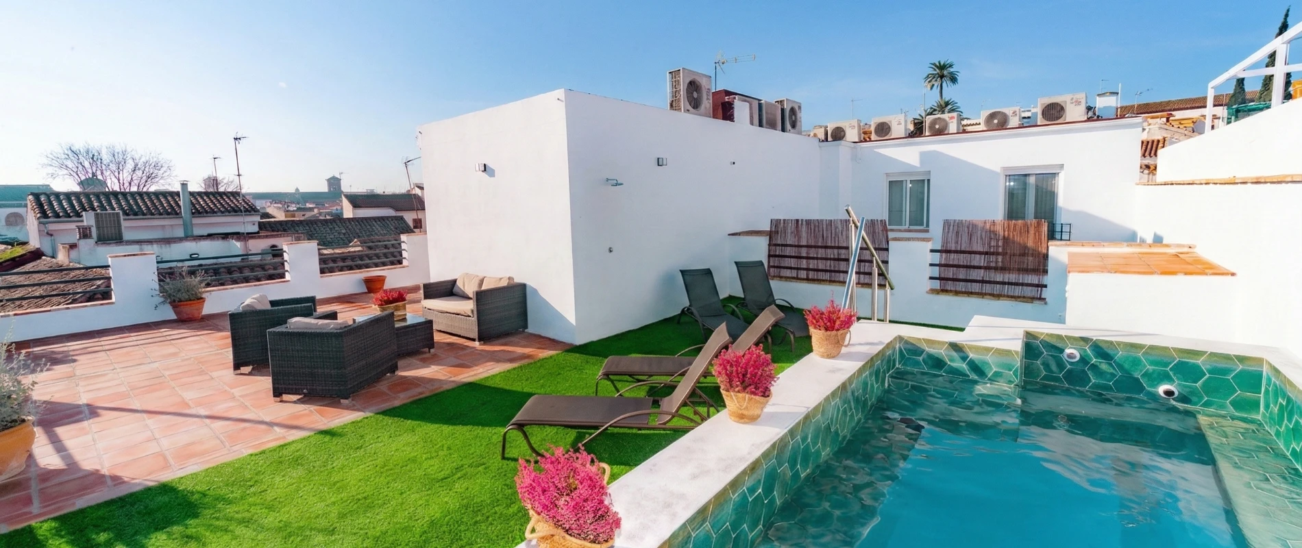 Terraza con piscina, tumbonas, sillones y plantas, rodeada de edificios bajo un cielo despejado.