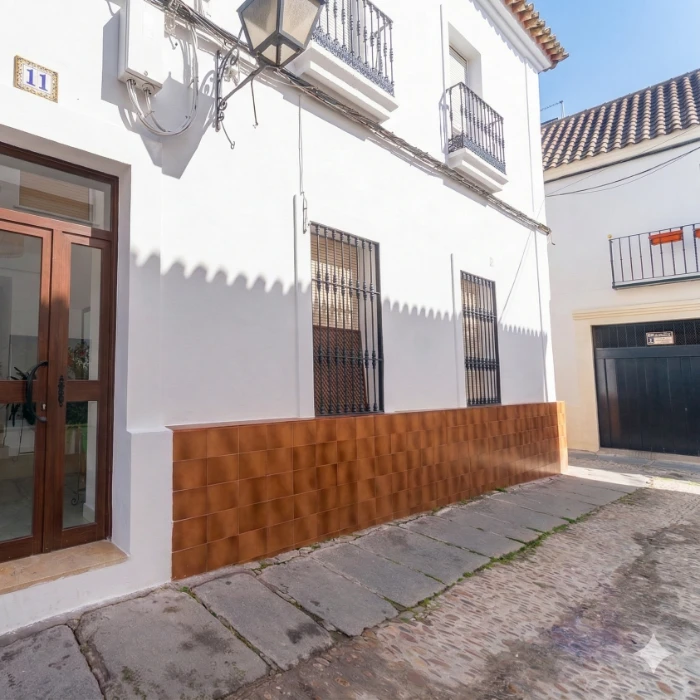 Fachada de una casa blanca con detalles en marr&oacute;n, puerta de madera y ventanas con rejas.