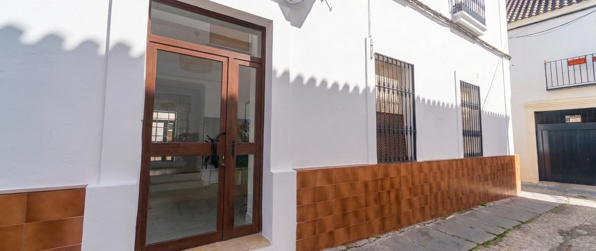 Edificio blanco con puerta de madera y ventanas con rejas, bajo luz solar directa.