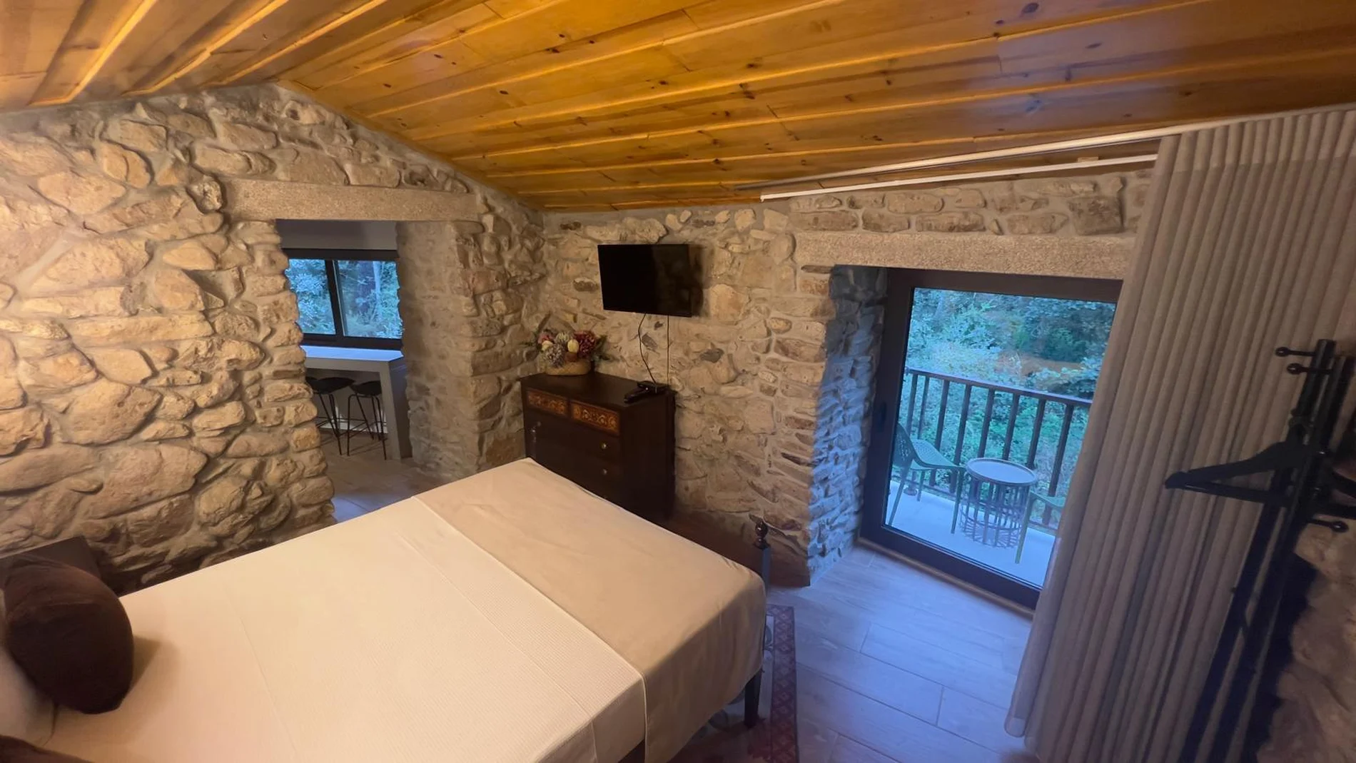 Quarto r&uacute;stico de pedra com cama, TV, varanda e vista para a natureza.