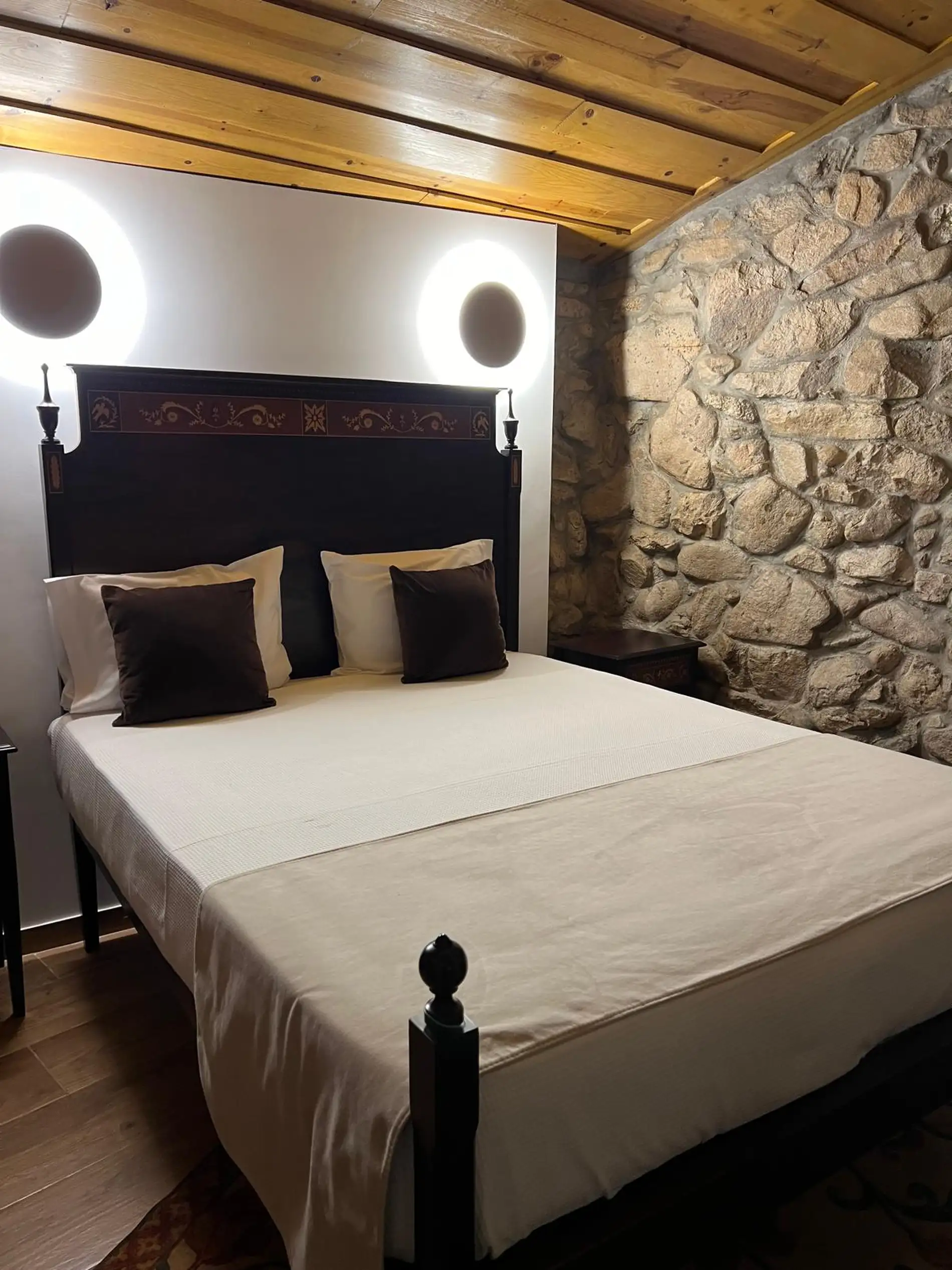 Cama de casal com cabeceira escura, luzes circulares e parede de pedra.
