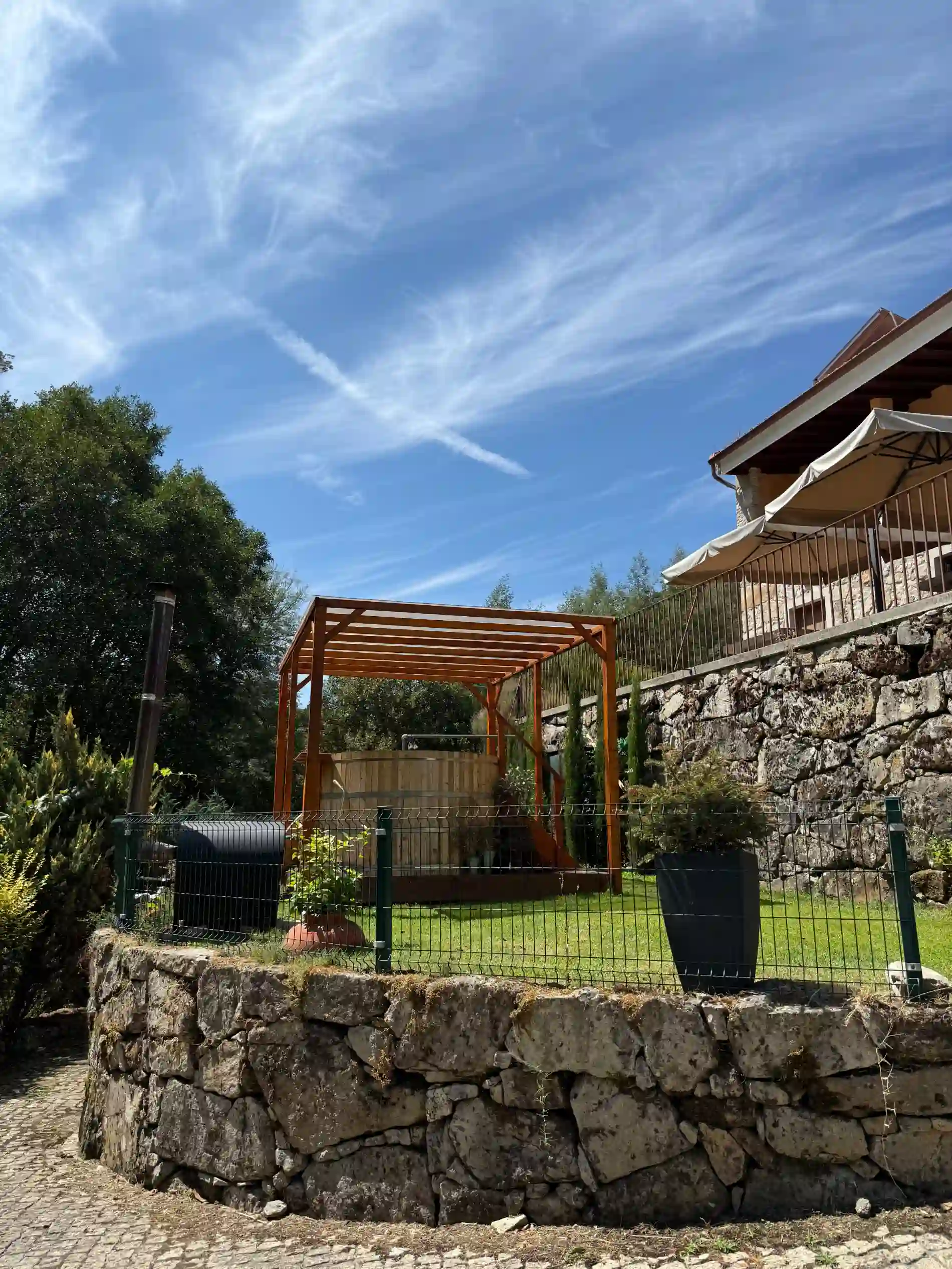 Casa de pedra com jardim e estrutura de madeira sob c&eacute;u azul.