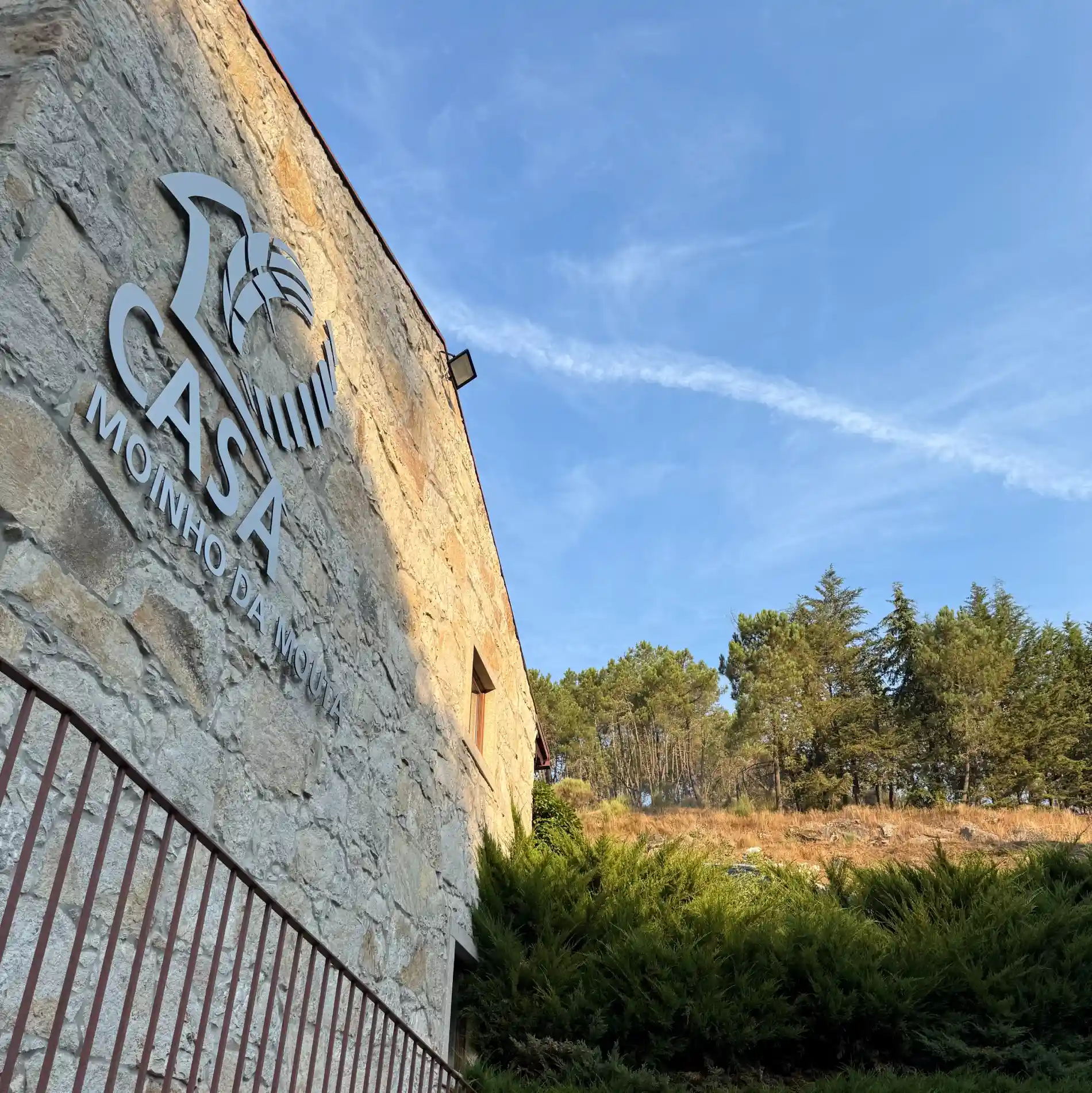 Parede de pedra com placa "Casa Moinho da Mouta", &aacute;rvores ao fundo.