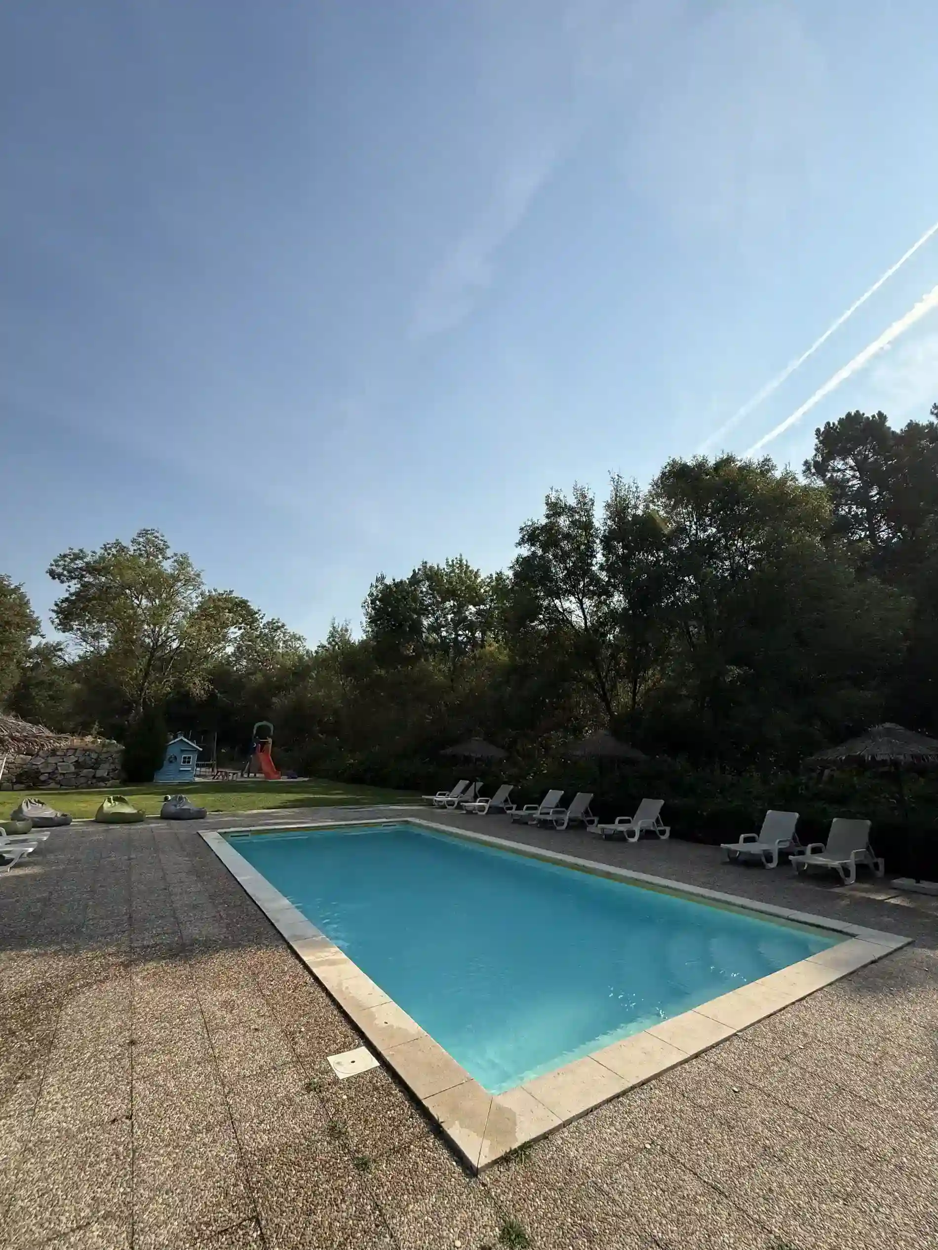 Piscina ao ar livre cercada por espregui&ccedil;adeiras e &aacute;rvores sob c&eacute;u azul.
