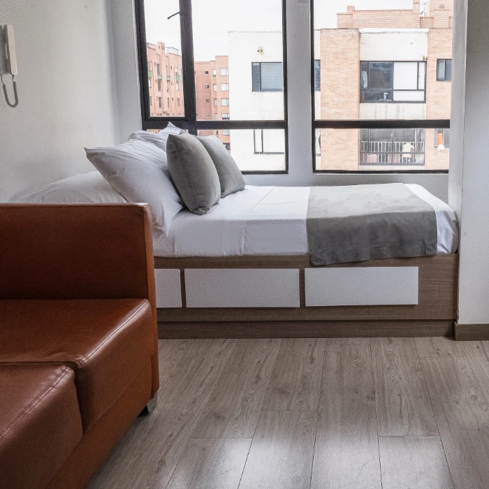 Habitaci&oacute;n minimalista con cama, sof&aacute; marr&oacute;n y ventana grande.