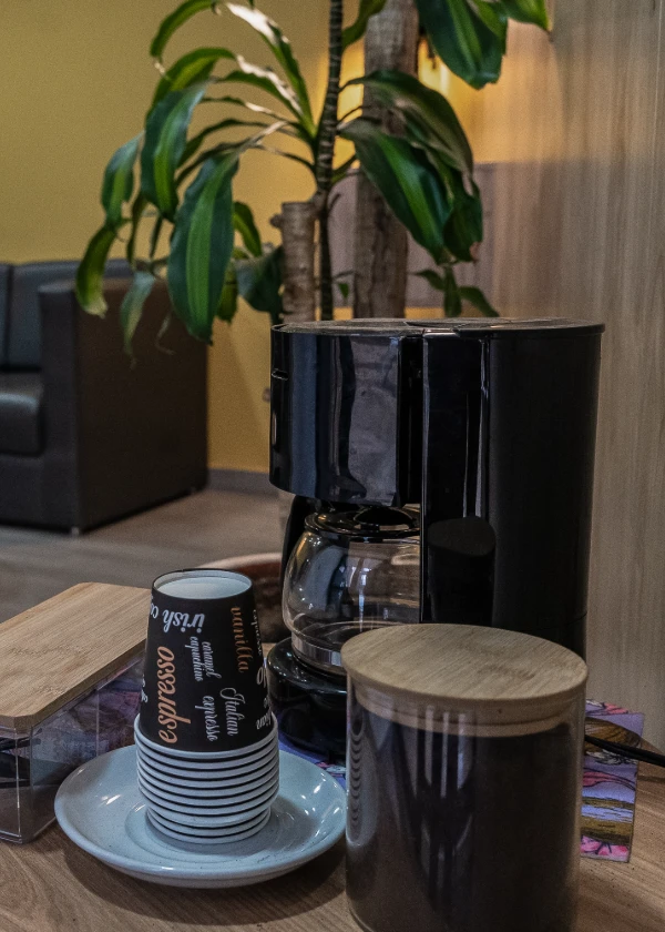 Cafetera negra con tazas y contenedores en una mesa, planta al fondo.