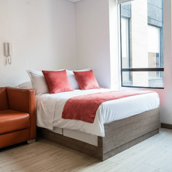 Habitaci&oacute;n moderna con cama y sof&aacute; naranja junto a una ventana grande.