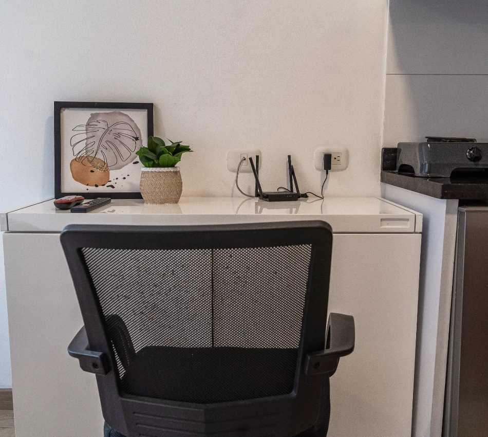 Oficina con escritorio blanco, silla negra, planta y cuadro decorativo.