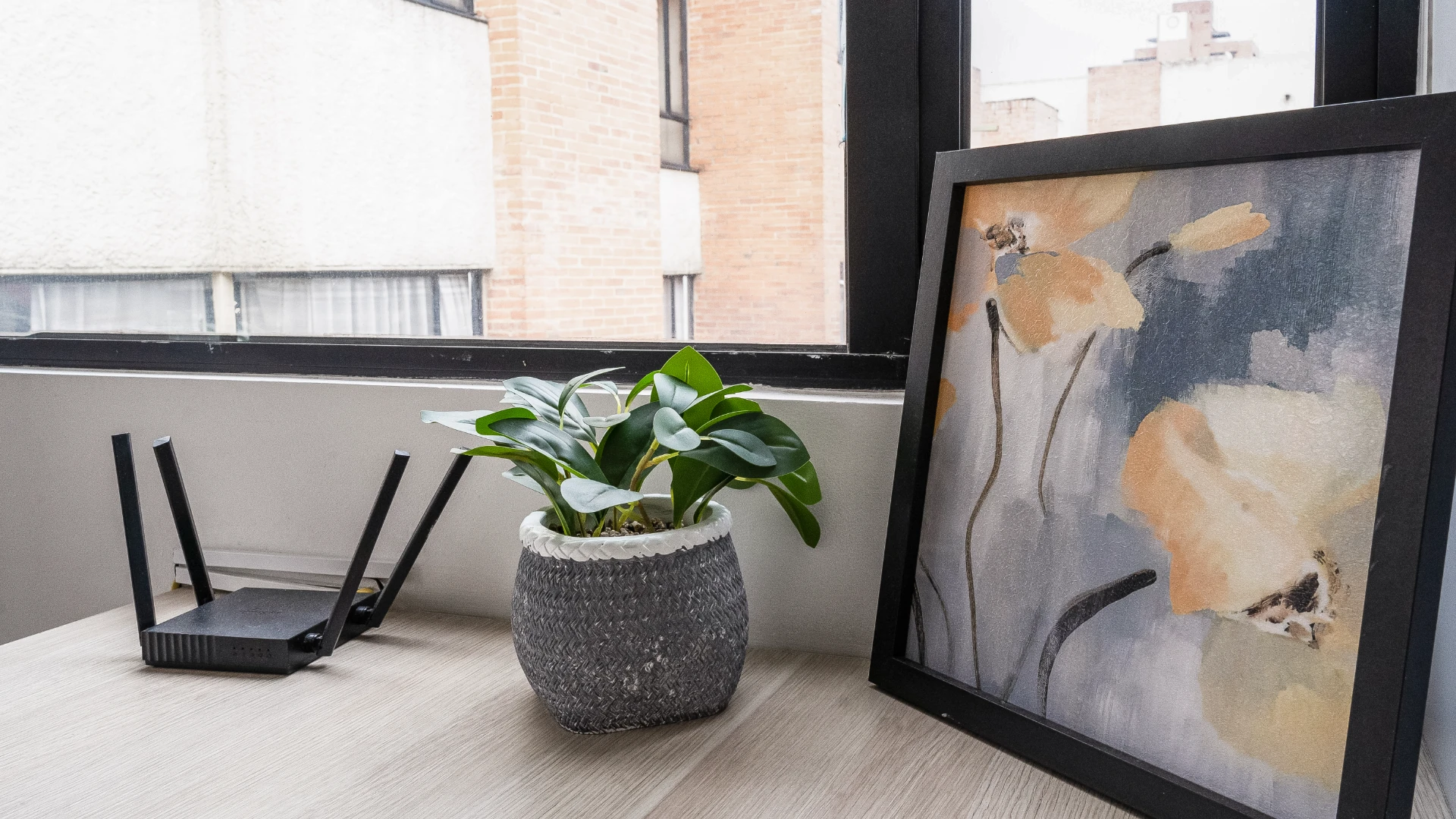 Planta, cuadro y router sobre mesa junto a una ventana con edificios al fondo.